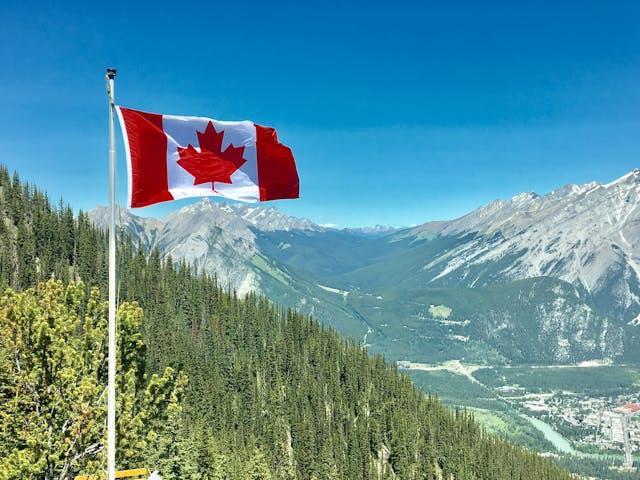 A Canadian flag on top of a flagpole, with mountains and forests in the background