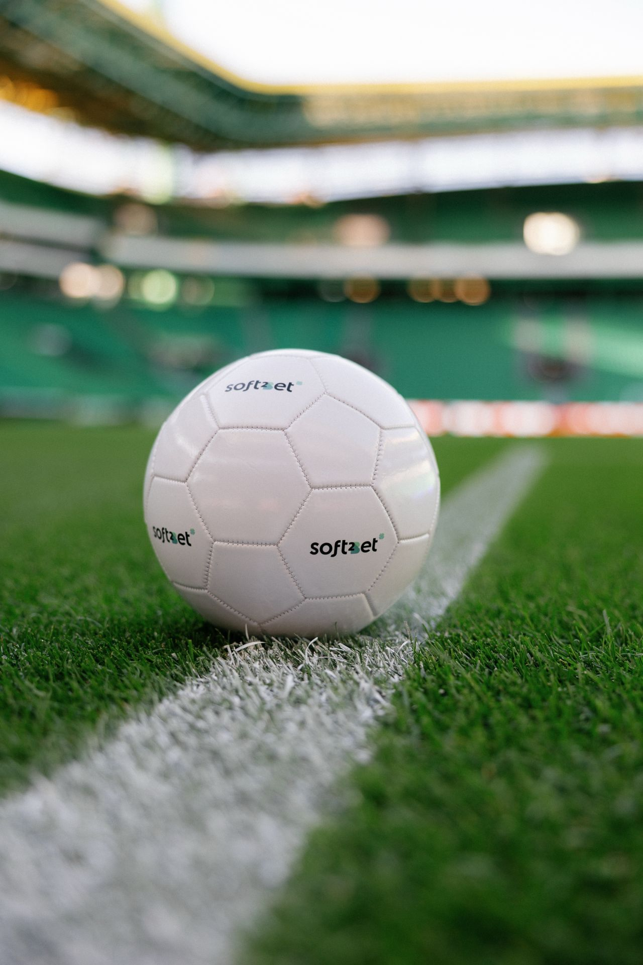 White football with Soft2Bet logo on a green stadium field, placed on the center line.