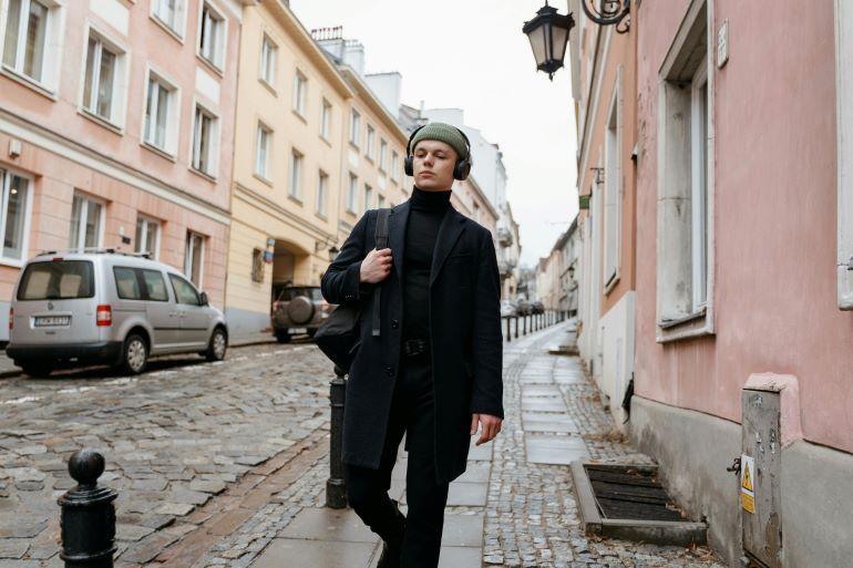 Person in a dark coat and green hat walks down a cobblestone street with headphones, listening to music.