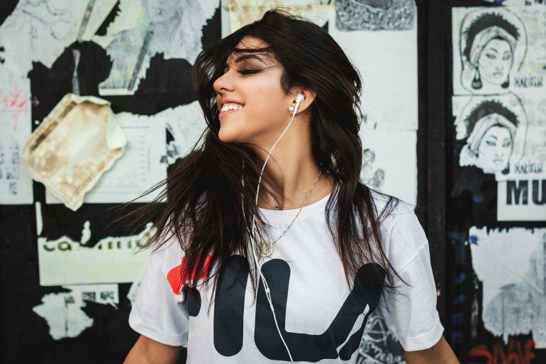 Young woman in a white T-shirt listens to music with earphones and smiles while moving joyfully outdoors.