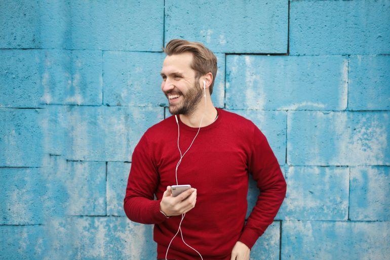 Man in a red sweater listens to music with earphones and smiles while holding a phone near a blue wall.