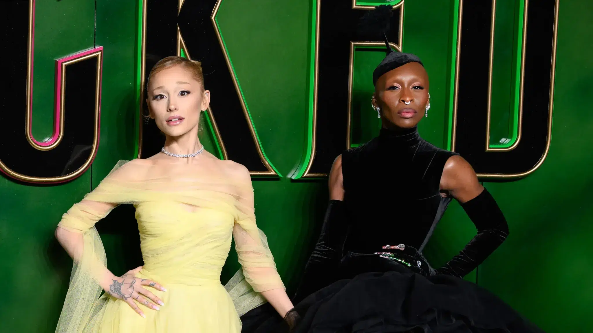 Ariana Grande and Cynthia Erivo in yellow and black dresses pose in front of a large Wicked backdrop