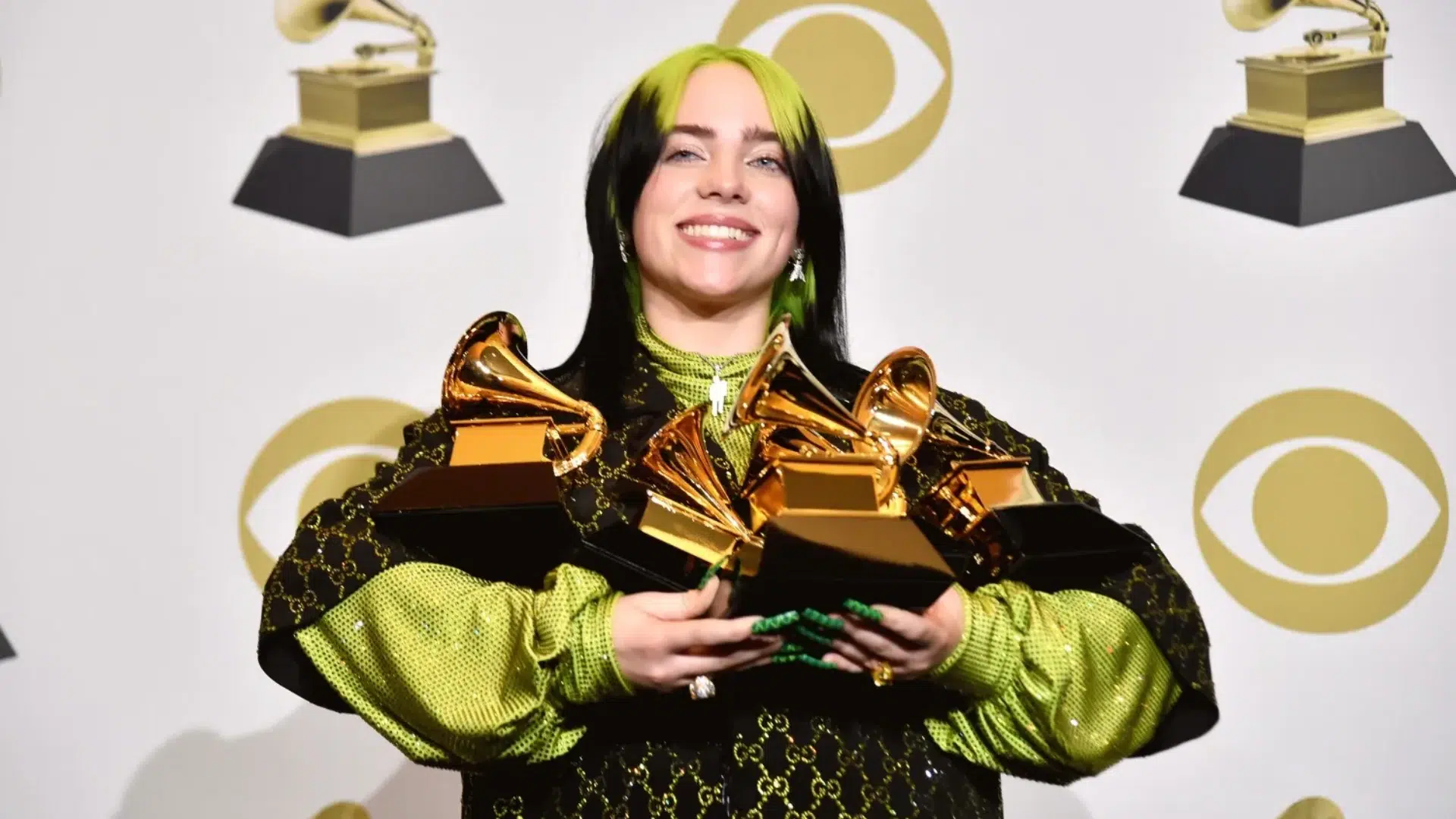 Billie Eilish smiles while holding multiple golden Grammy awards backstage, featuring her signature black and neon green hair and outfit