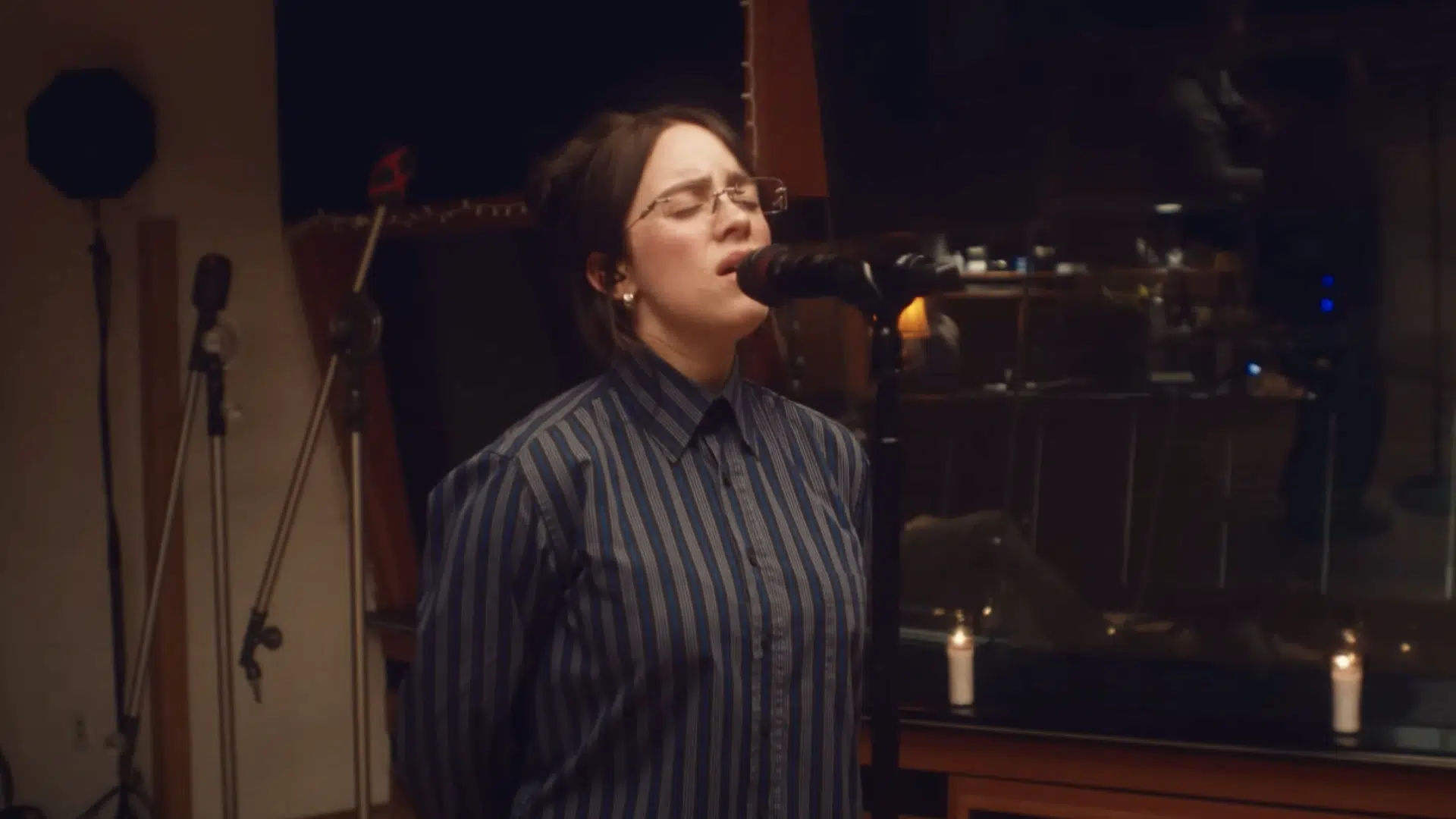 Billie Eilish, wearing glasses and a striped button-down shirt, is singing Wildflower into a microphone in a dimly lit recording studio.
