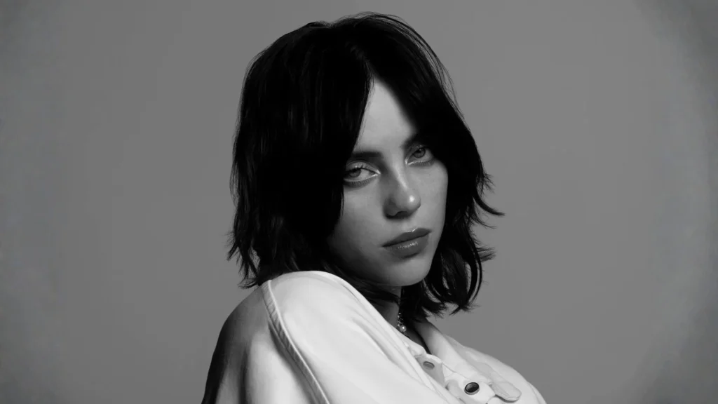 Black and white close-up portrait of Billie Eilish with dark, shaggy hair looking intently at the camera against a neutral gray background