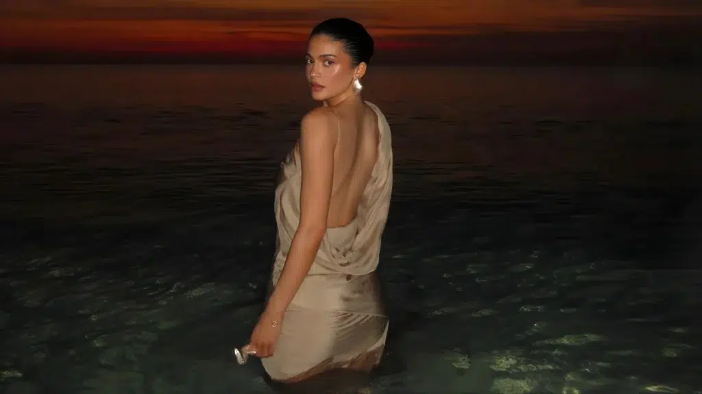 Kylie Jenner standing in shallow water at sunset wearing a backless beige dress and looking over her shoulder.