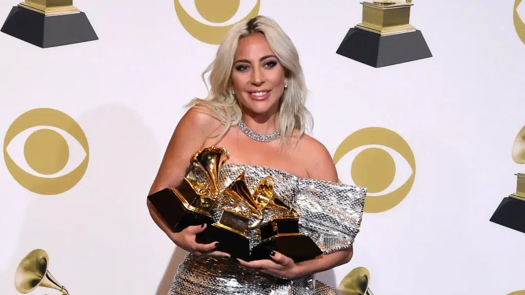 Lady Gaga holding multiple gold Grammy awards in front of a white backdrop featuring the CBS eye logo