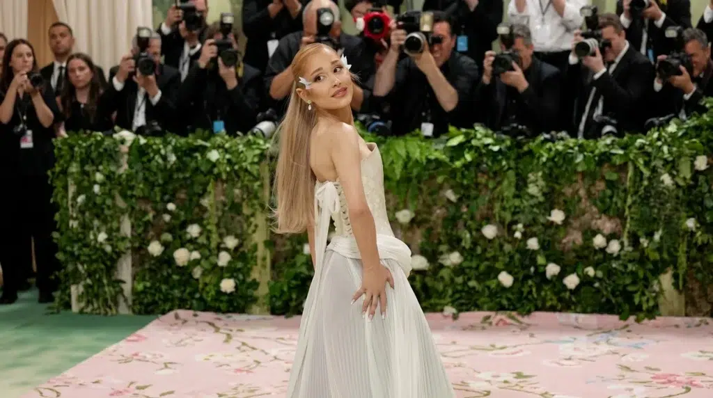 ariana grande at the met gala wearing a white corset dress, posing for photographers with a floral backdrop and a smile