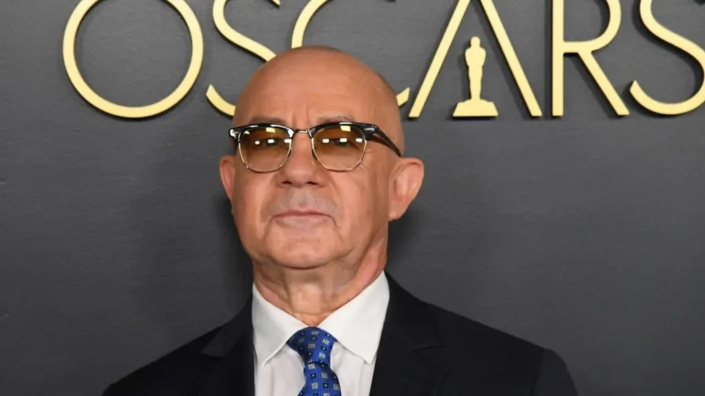 bernie taupin wearing glasses and a suit with a blue patterned tie stands in front of a black wall with the word oscars
