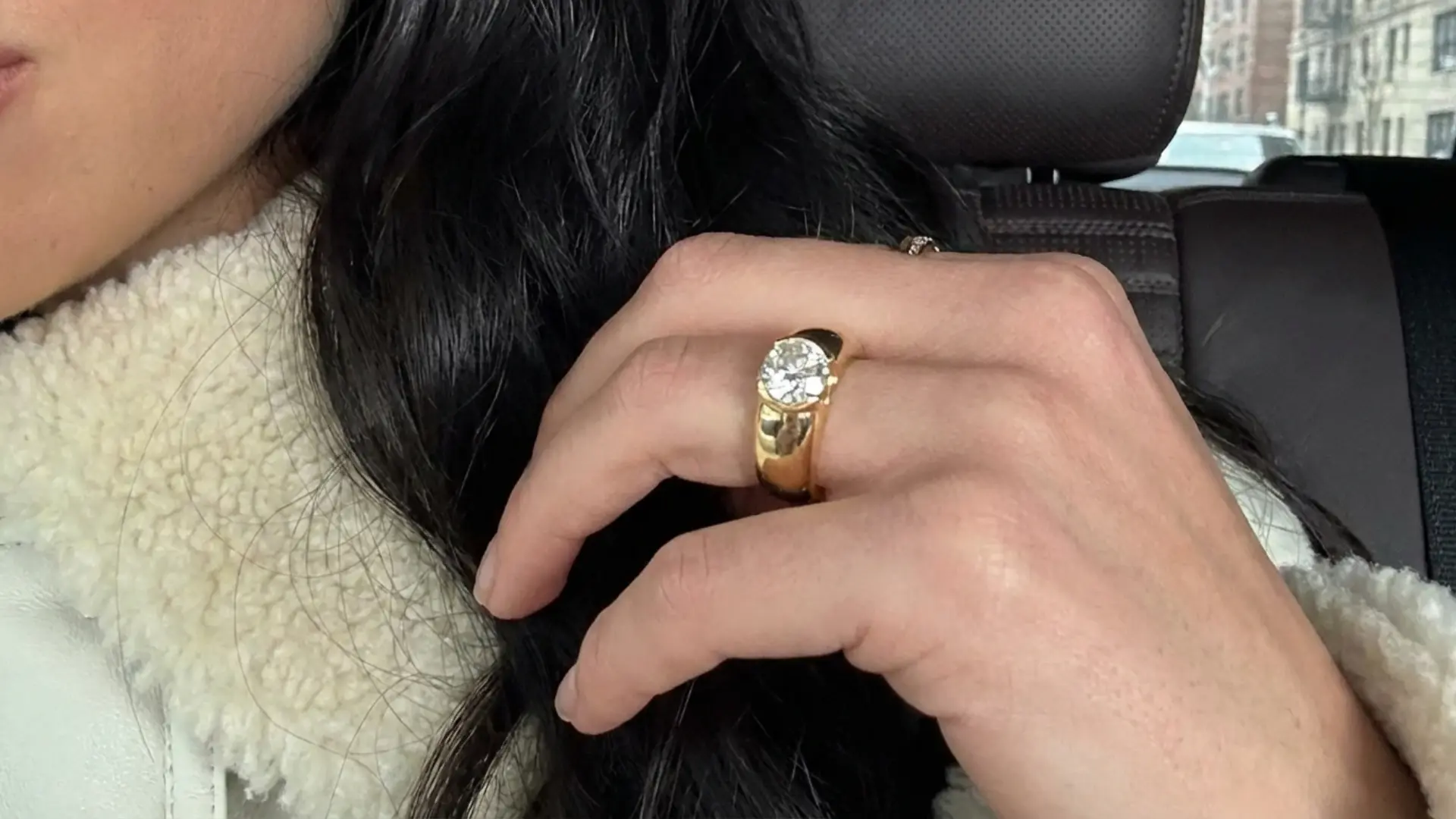 close-up of a hand with a thick gold ring featuring a large diamond while wearing a white sherpa-lined jacket