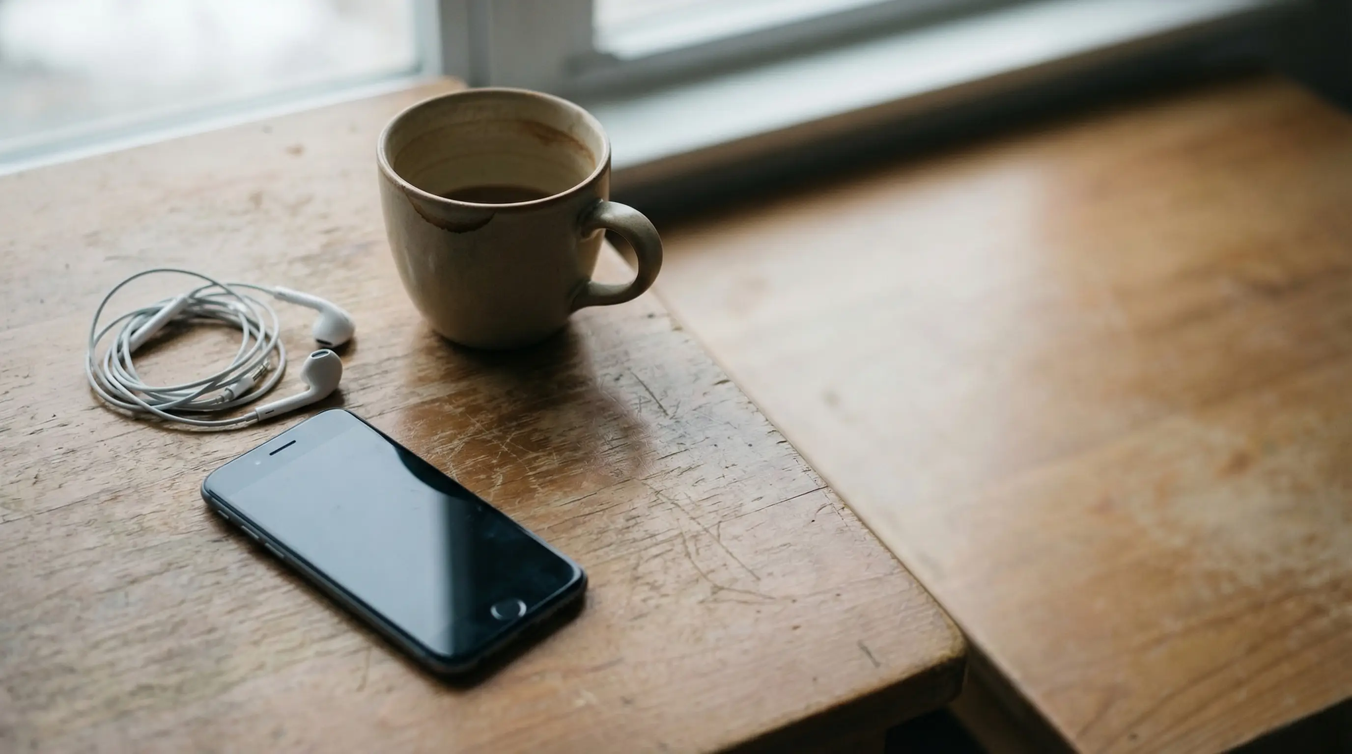 Smartphone with earphones and coffee mug on wooden table near window