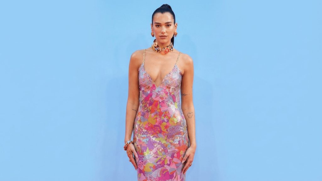 dua lipa wearing a colorful floral sequin slip dress with gold chain straps and statement jewelry against a blue background