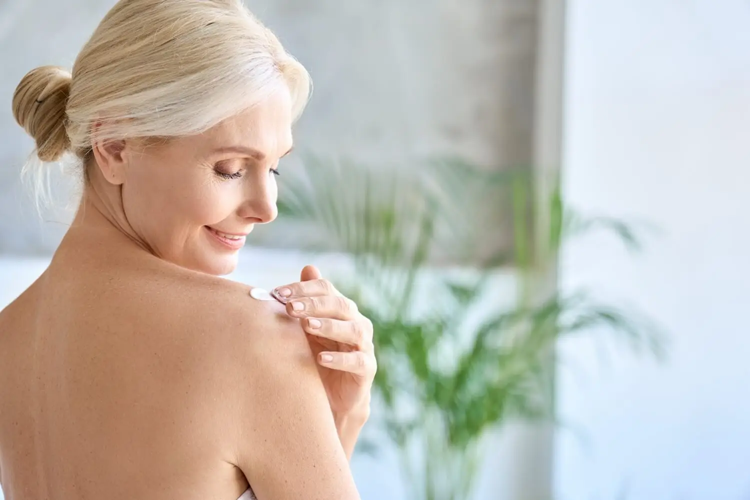 Woman applying cream on shoulder in bright, indoor setting with blurred plant background