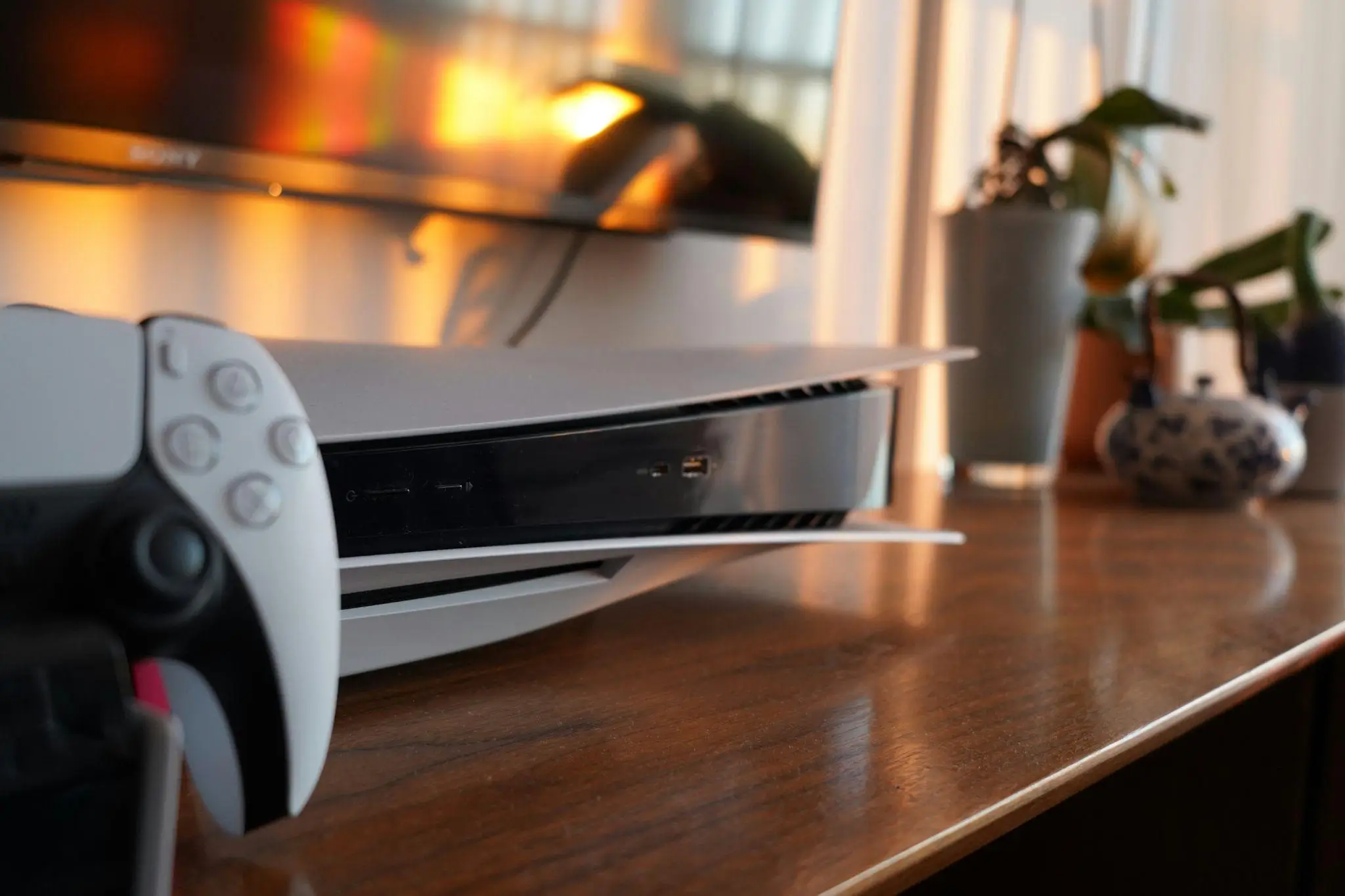 Gaming console and controller on wooden media unit with potted plants in warm ambient lighting