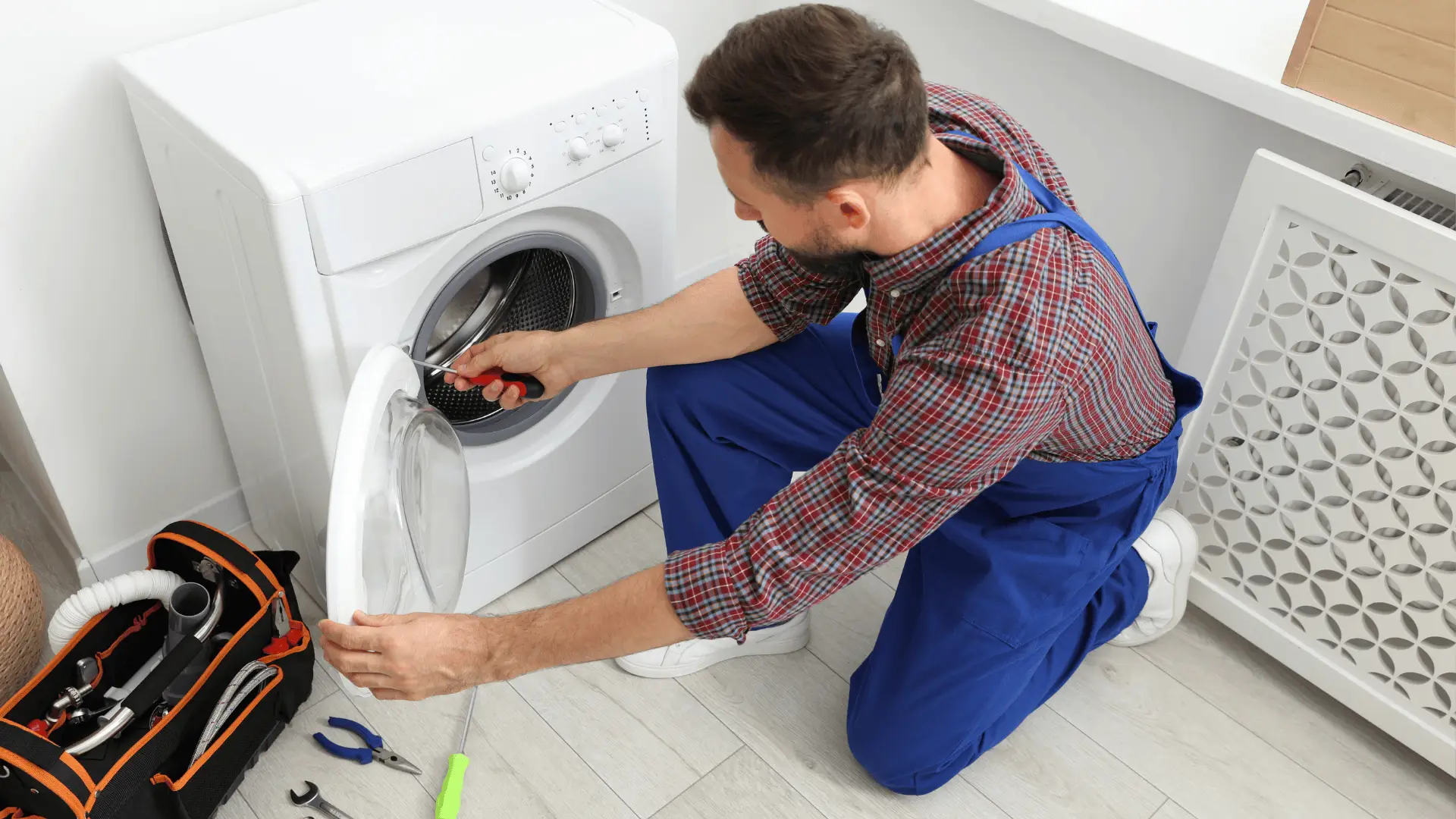Repairman fixing washing machine with screwdriver in laundry room, tool bag on floor