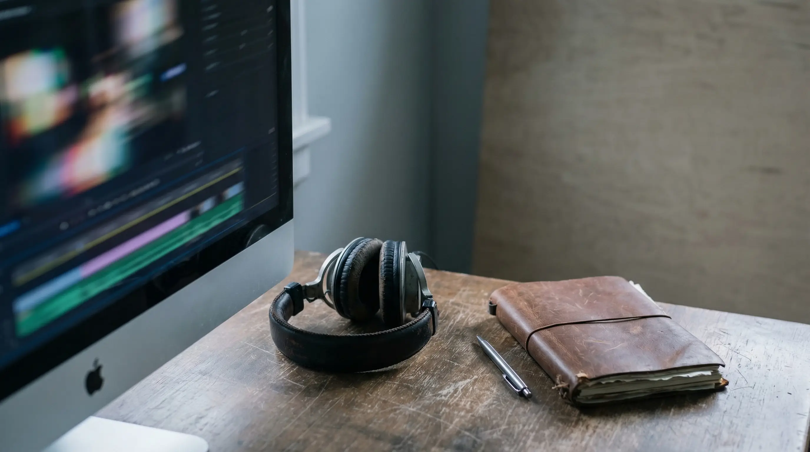 Computer monitor displaying video editing software with headphones, notebook, and pen on wooden desk