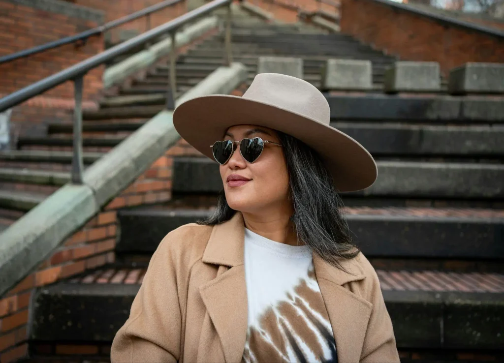 Woman in tan coat and wide-brimmed hat standing on brick staircase wearing heart-shaped sunglasses