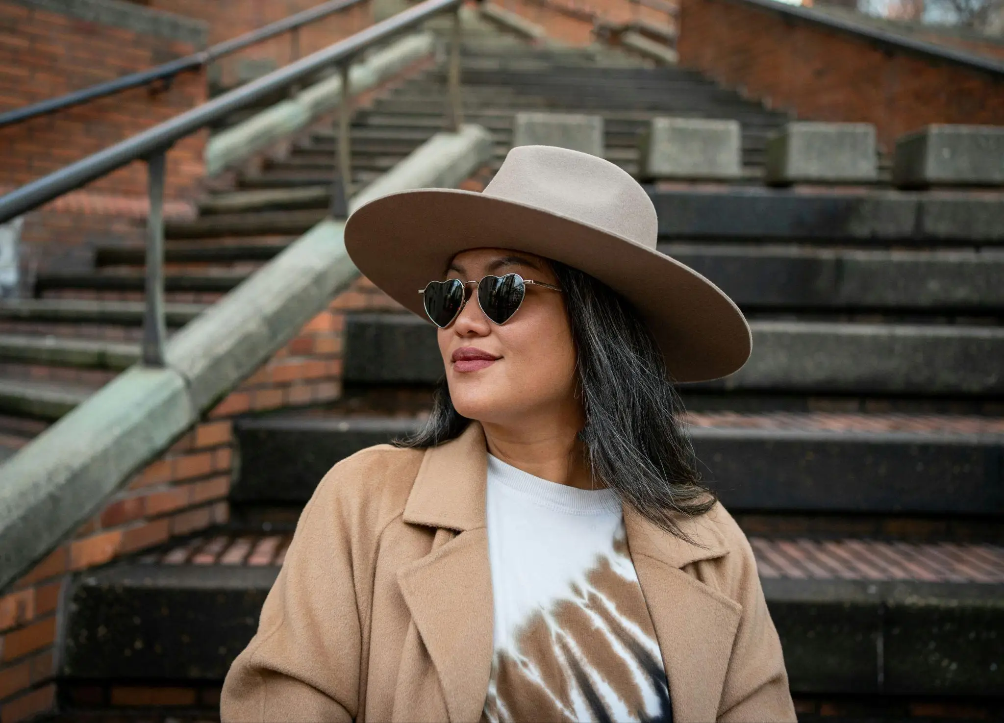 Woman in tan coat and wide-brimmed hat standing on brick staircase wearing heart-shaped sunglasses