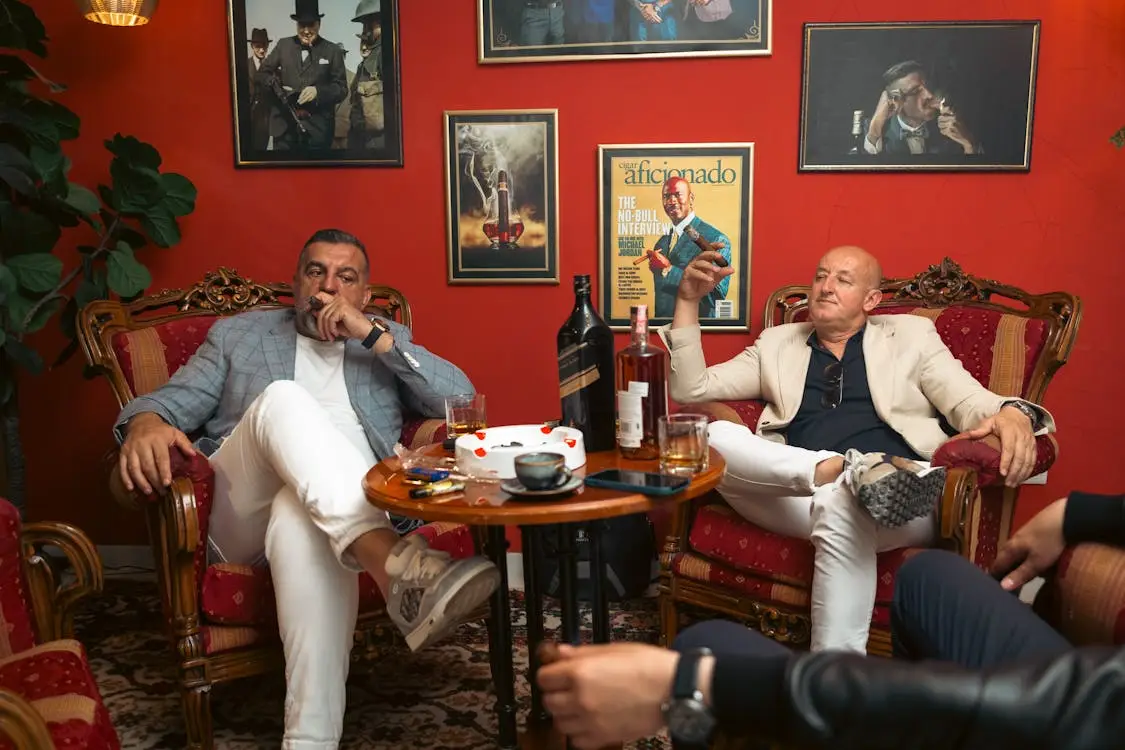 Two men sitting in armchairs smoking cigars in a warmly lit lounge with red walls