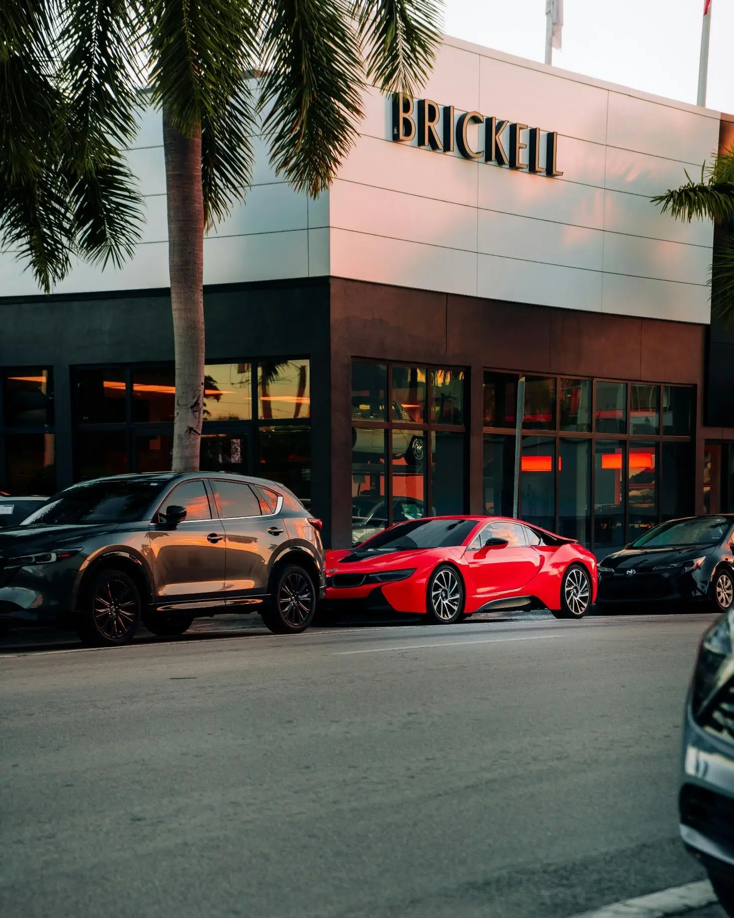 Red sports car and grey SUV parked outside modern building with large windows and palm trees