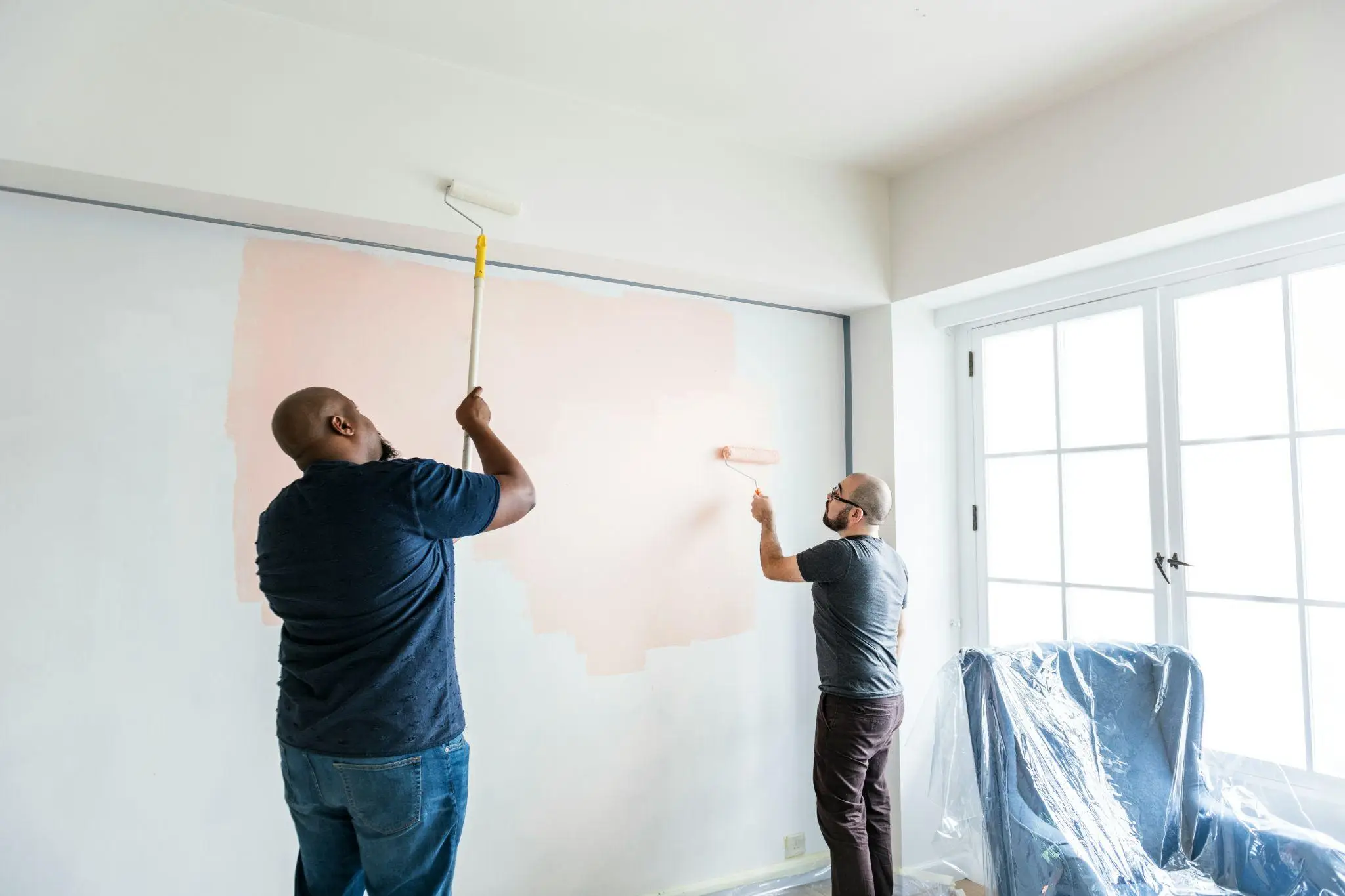 Two men painting a beige wall in a bright living room with plastic-covered chair nearby