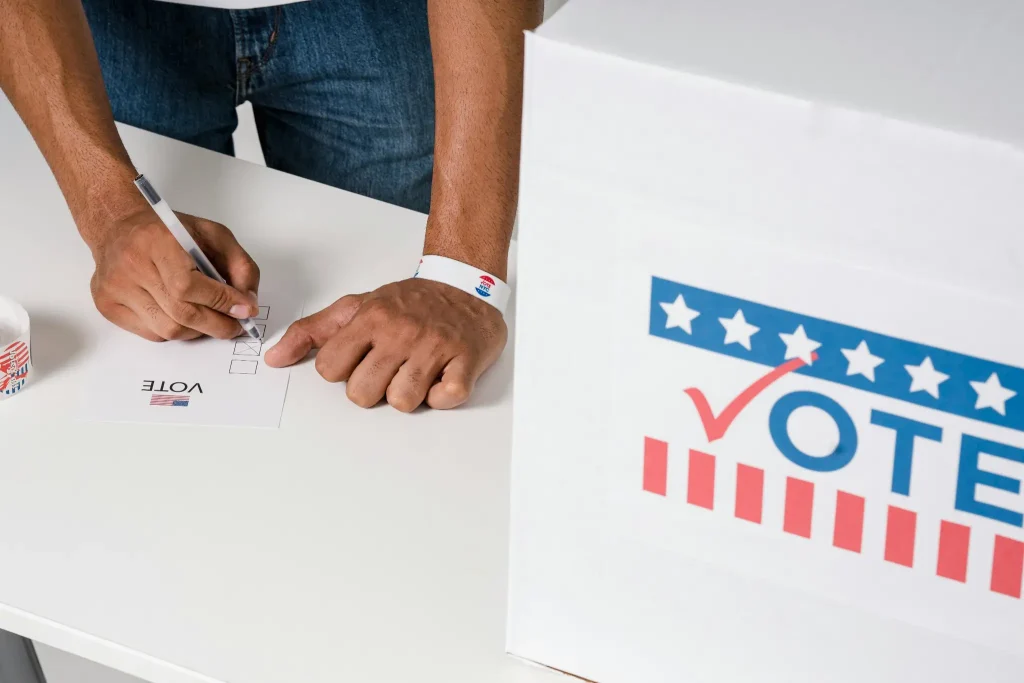 Person marking ballot paper on white table next to voting box with patriotic design