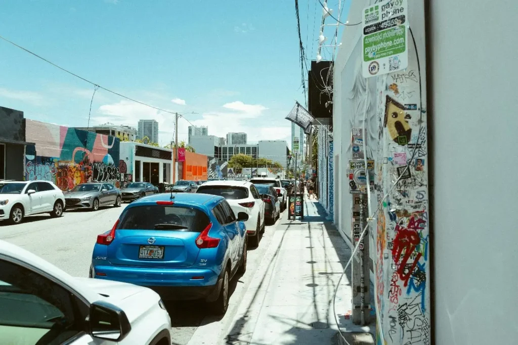 Street lined with parked cars, colorful murals, and graffiti-covered walls in urban setting
