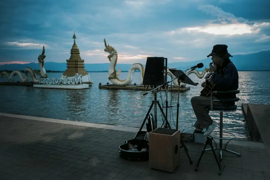 Musician playing guitar by waterfront at sunset with ornamental dragon statues in the background