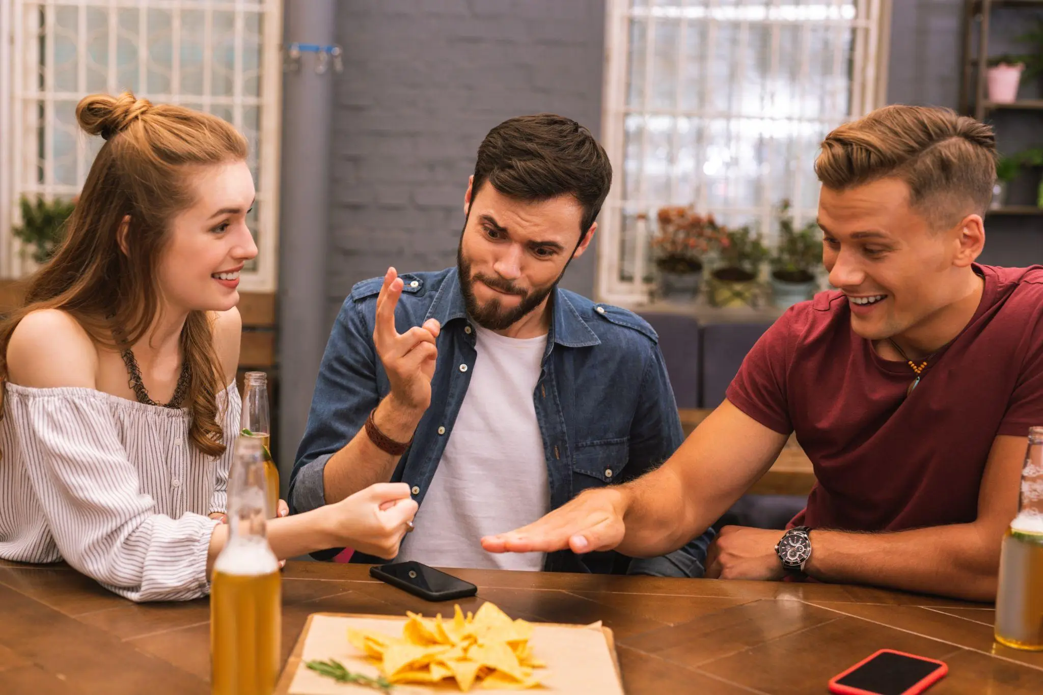 Three friends playing rock-paper-scissors at a wooden table with drinks and snacks