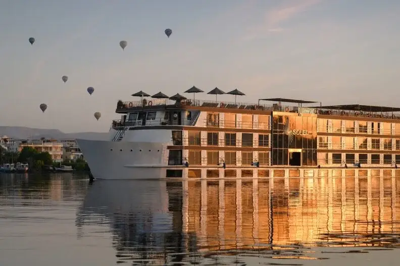 Large river cruise ship with multiple decks reflecting sunset light on calm water with hot air balloons overhead