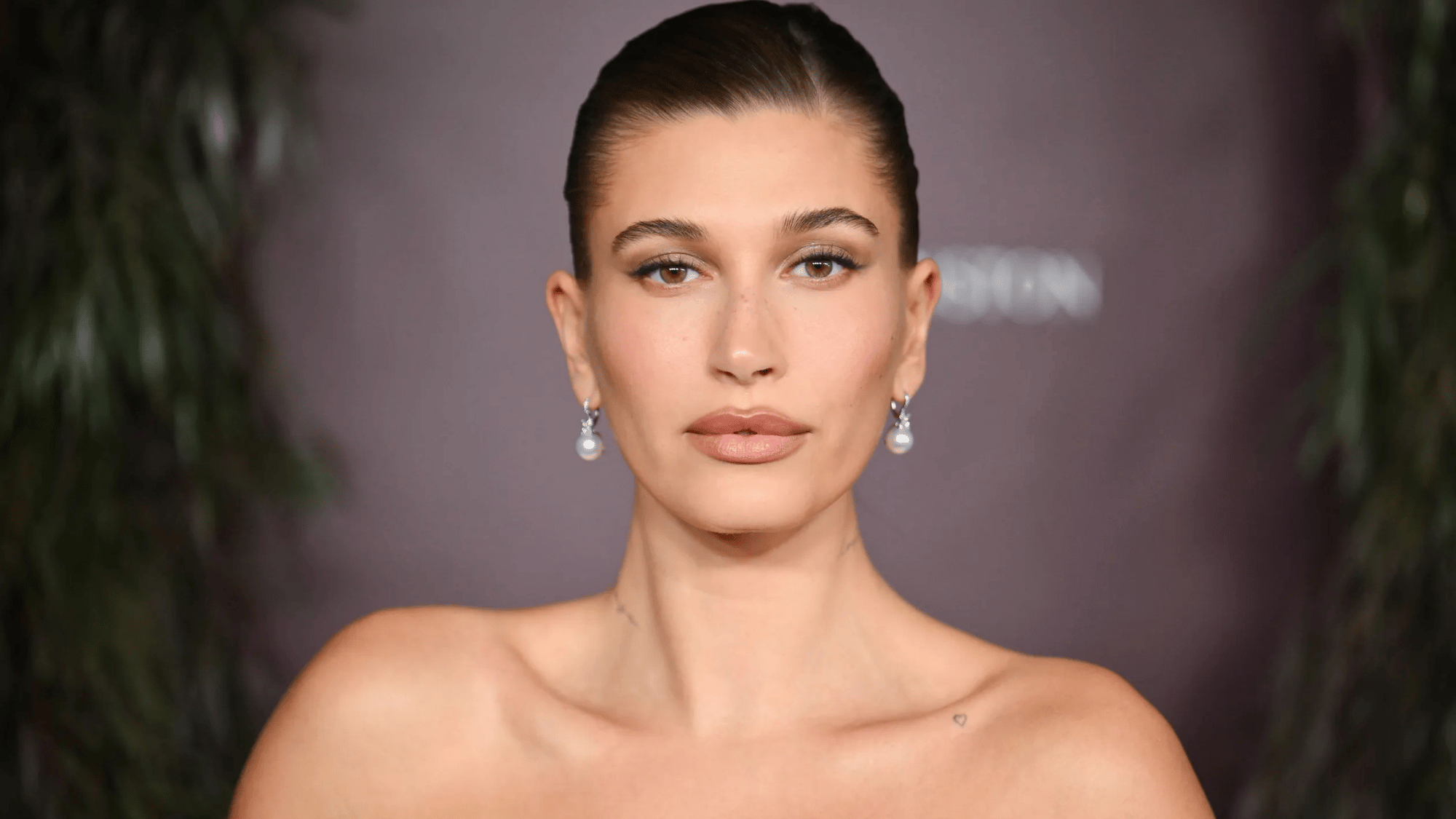 Hailey Bieber posing at a formal event with slicked-back hair, natural makeup, pearl drop earrings, and a neutral background