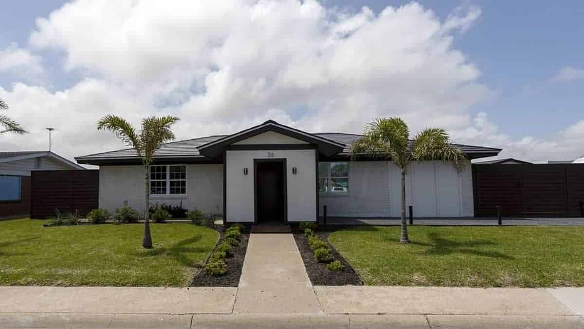 Modern single-story white house with black trim, palm-lined walkway, manicured lawn and cloudy sky overhead above