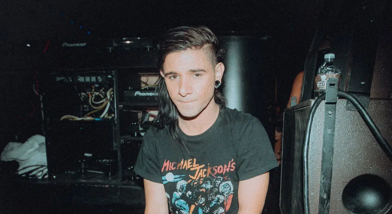 Skrillex sitting backstage near DJ equipment while wearing a graphic T-shirt, showing his casual style and electronic music background.
