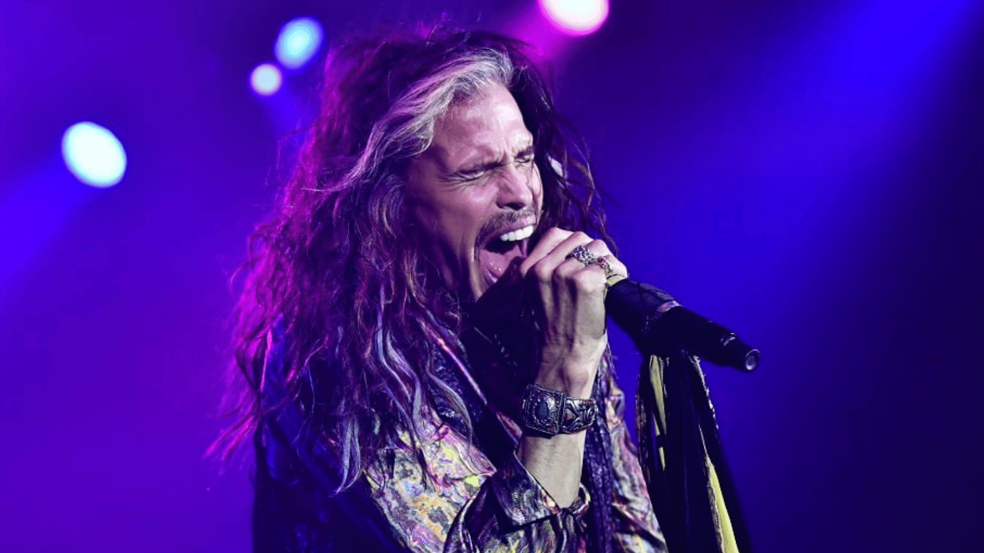 Steven Tyler passionately singing into a microphone on stage under bright purple concert lights during a live performance.