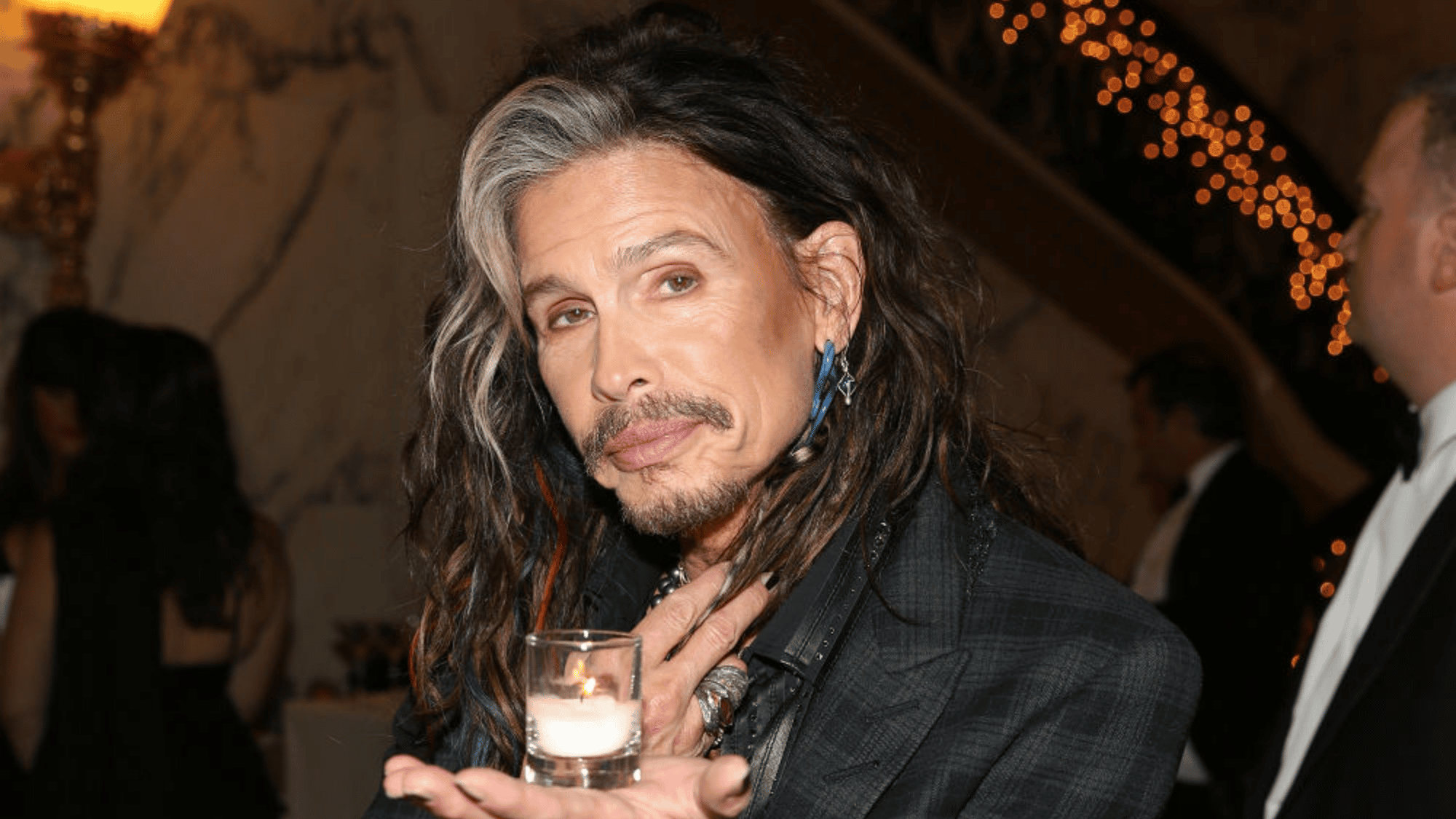 Steven Tyler posing at an upscale indoor event with long hair, gray streaks, and formal clothing while holding a lit candle.