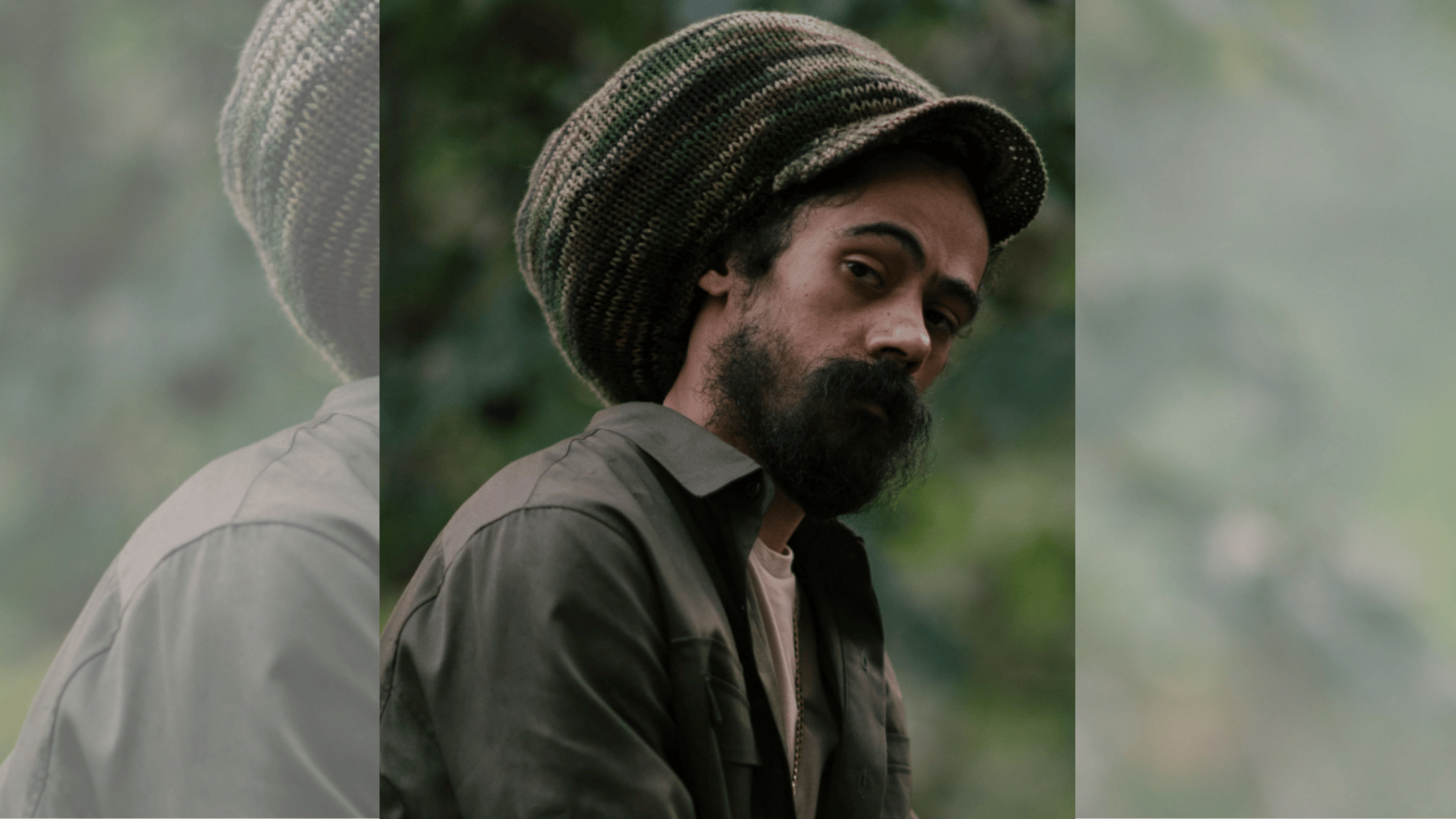 bearded man in oversized knit tam standing outdoors with blurred green background.