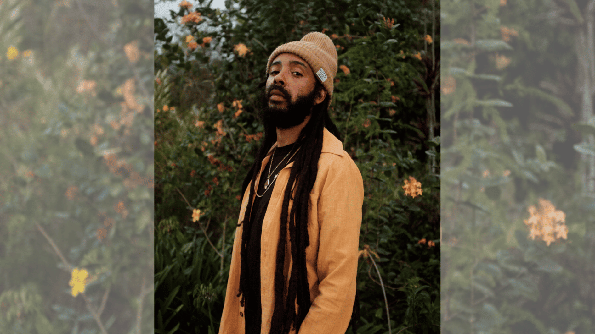 bearded reggae artist in tan beanie and mustard jacket standing among lush garden plants