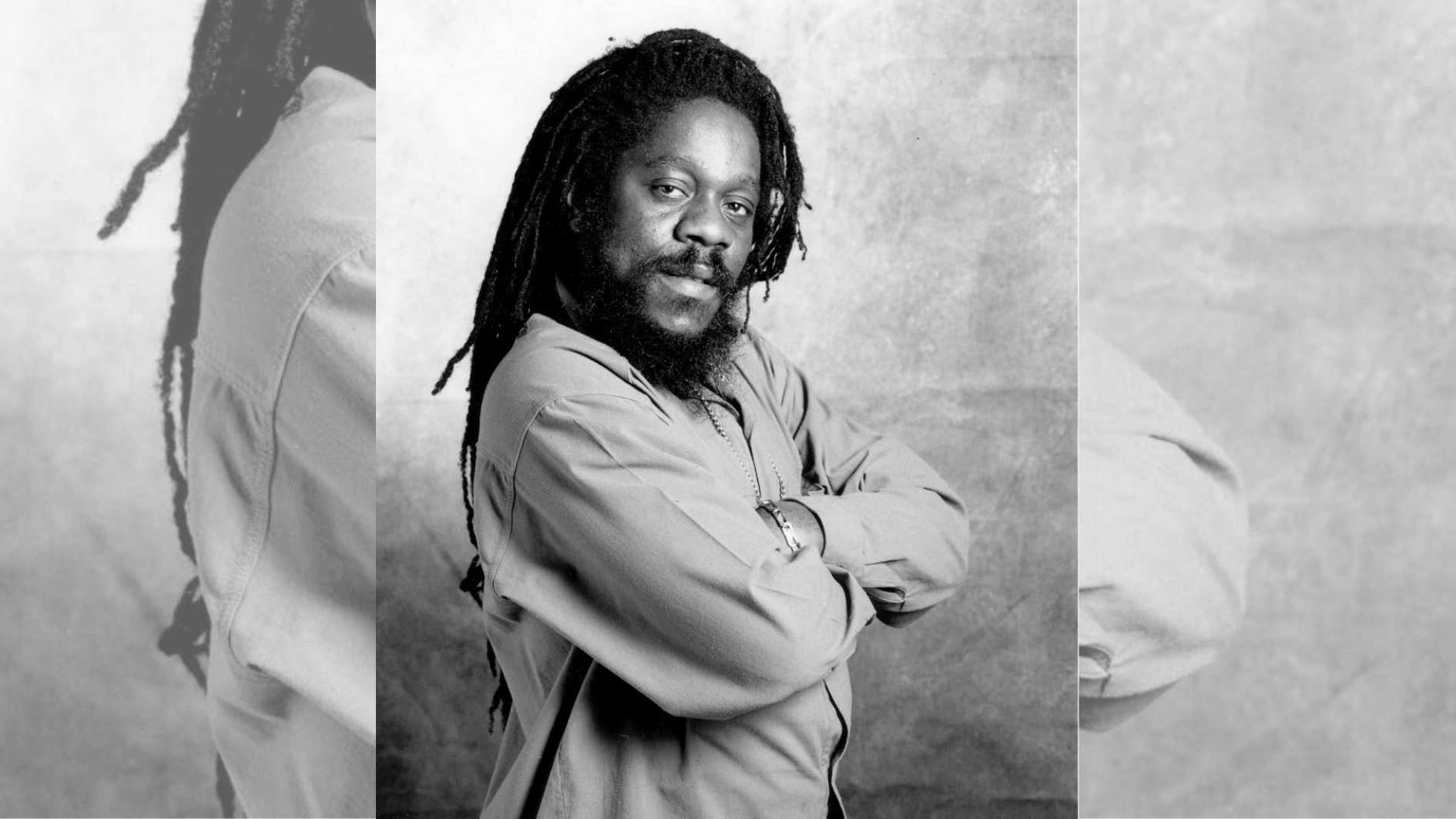 black-and-white portrait of man with dreadlocks posing with folded arms in loose shirt.