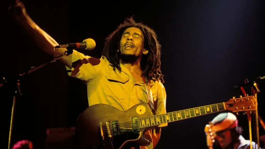 bob marley sings with his eyes closed while playing an electric guitar on stage under warm yellow lights