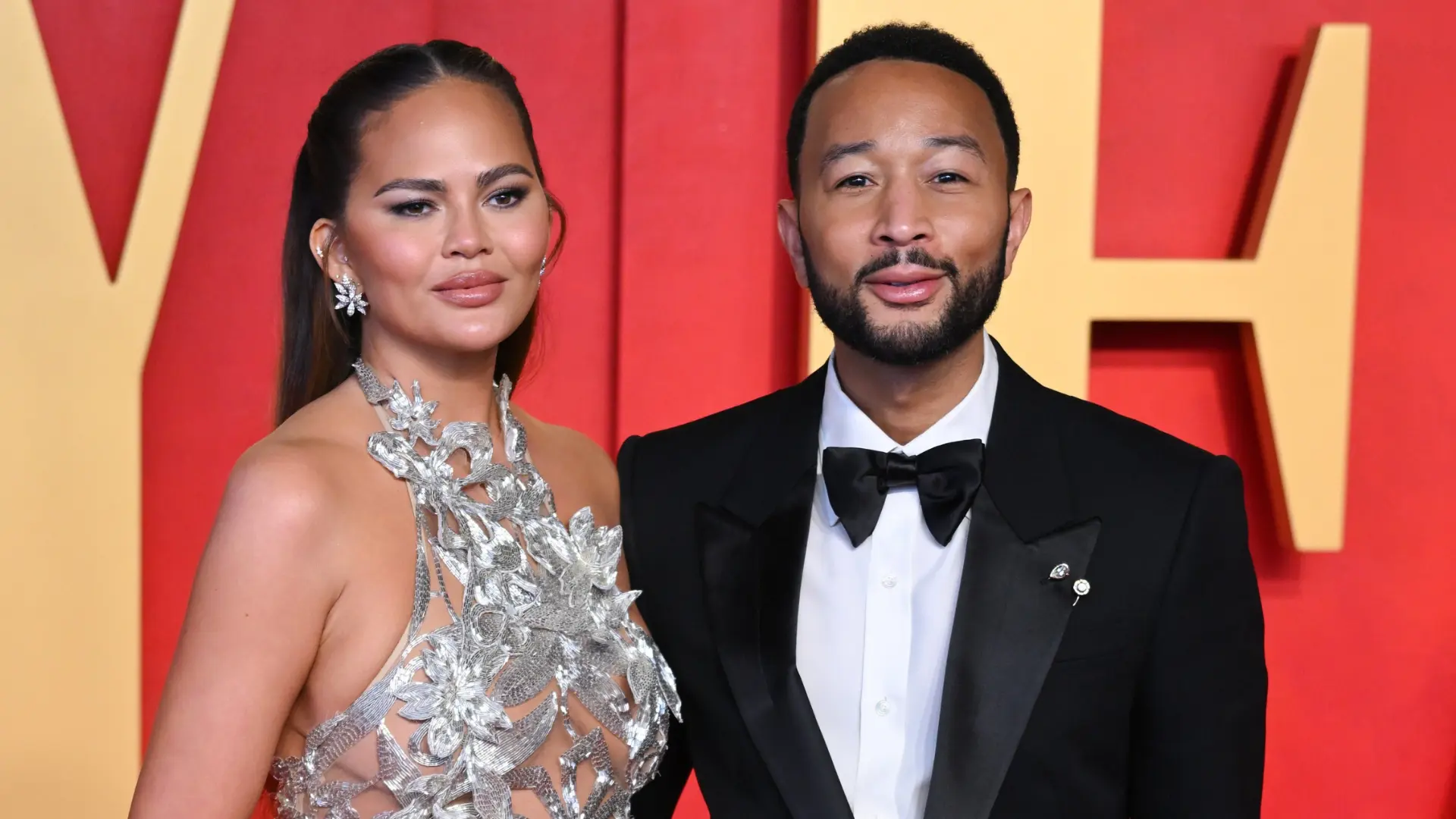 chrissy teigen in a silver floral dress and john legend in a tuxedo pose together against a red and gold background