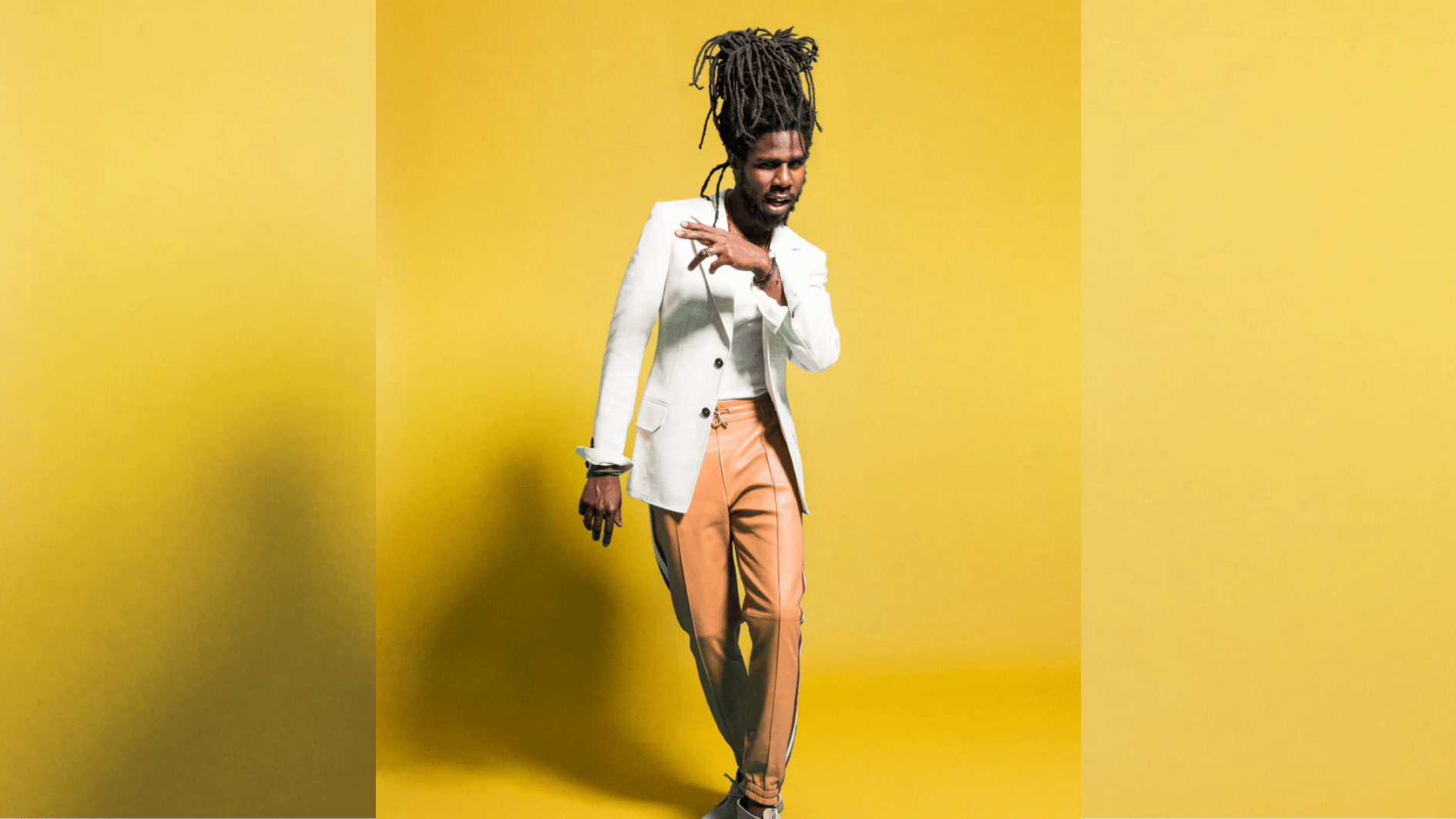 dreadlocked performer in white blazer and tan trousers posing against vivid yellow backdrop.