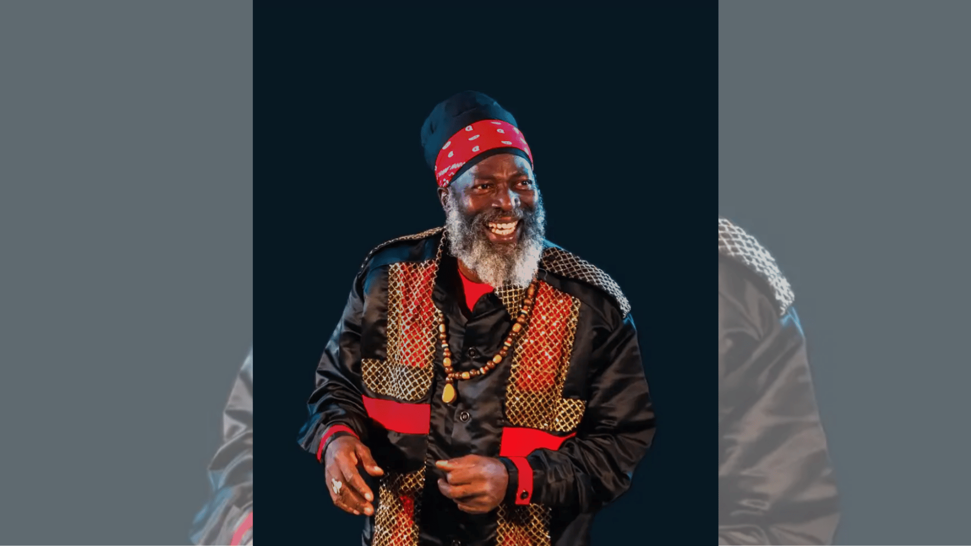 gray-bearded reggae artist in red headscarf smiling in black and gold stage outfit.