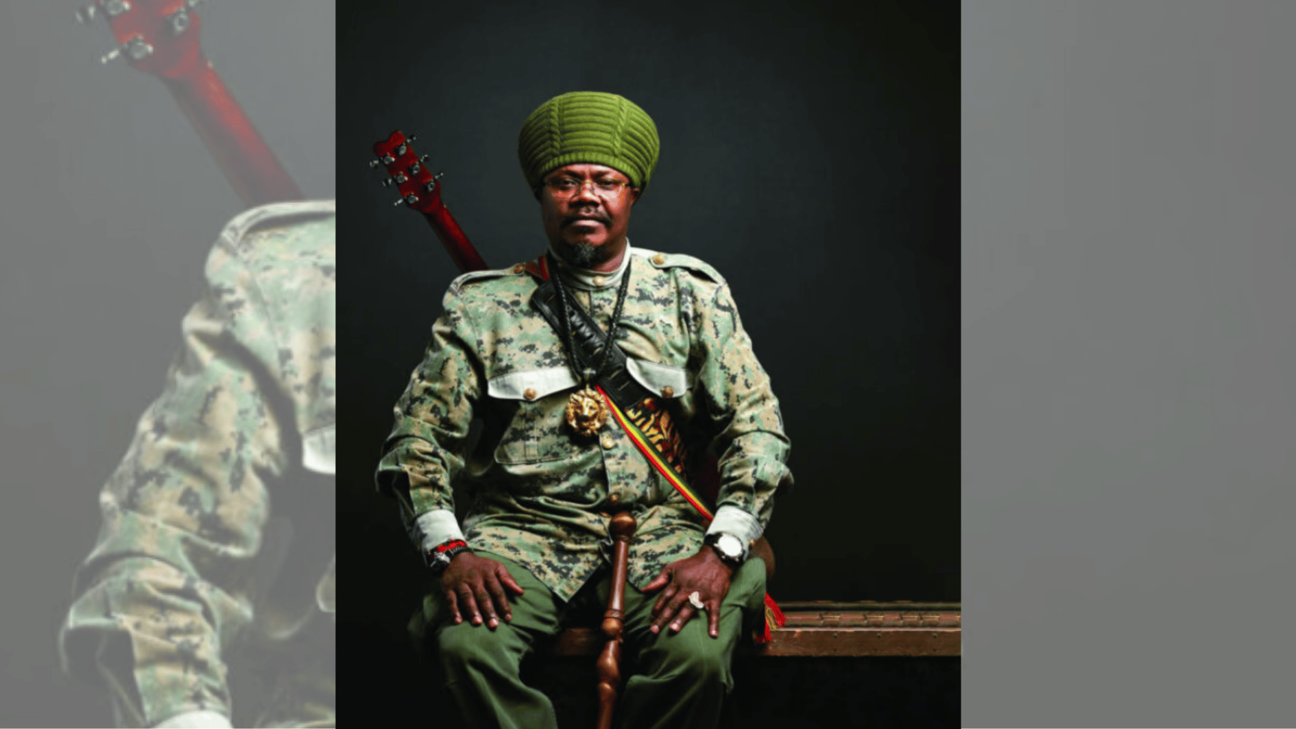 man in camouflage outfit and green knit turban seated with guitar against dark backdrop.