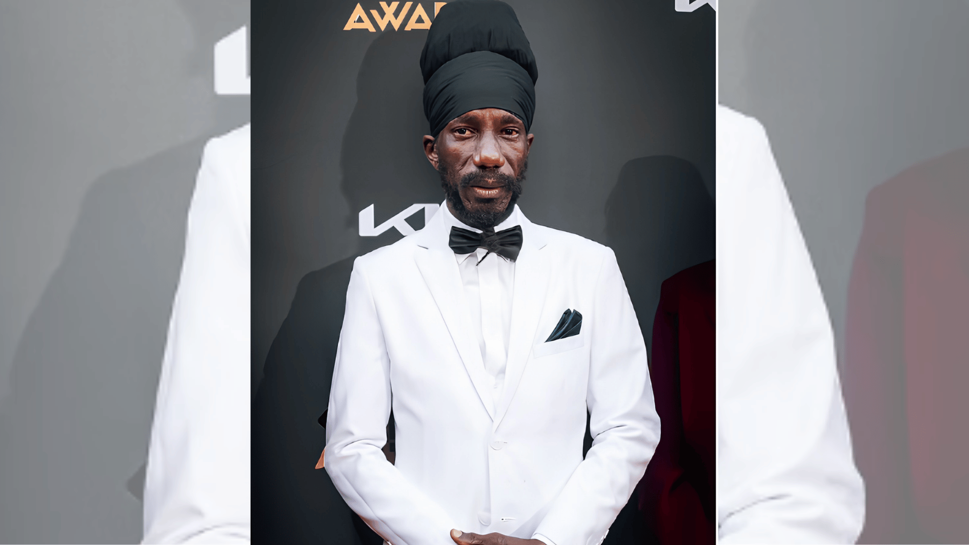 man in white tuxedo and tall black headwrap posing at a Caribbean music awards event.