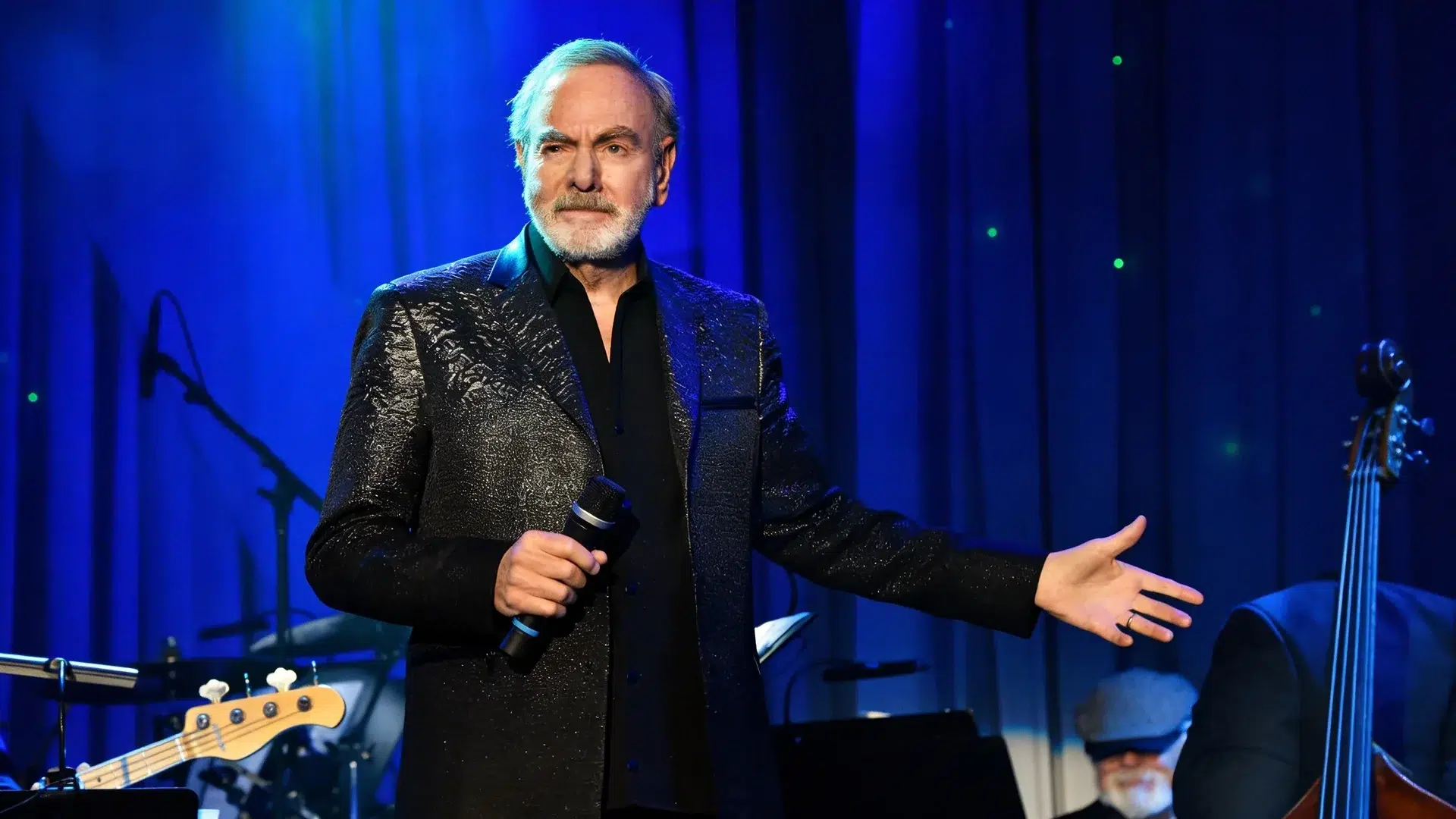 neil diamond performing live on stage, holding a microphone, wearing a dark jacket, with a band behind and blue lighting