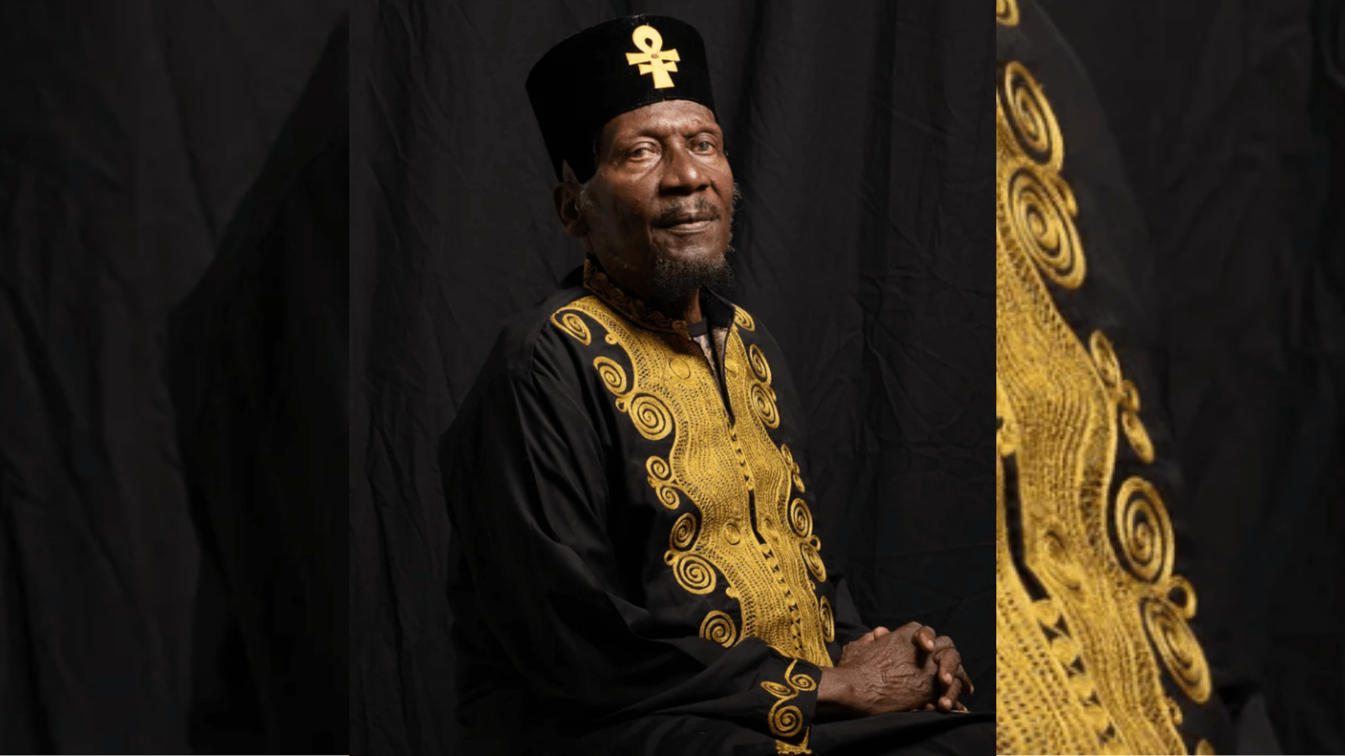older man in black robe with gold embroidery posing against dark studio backdrop.