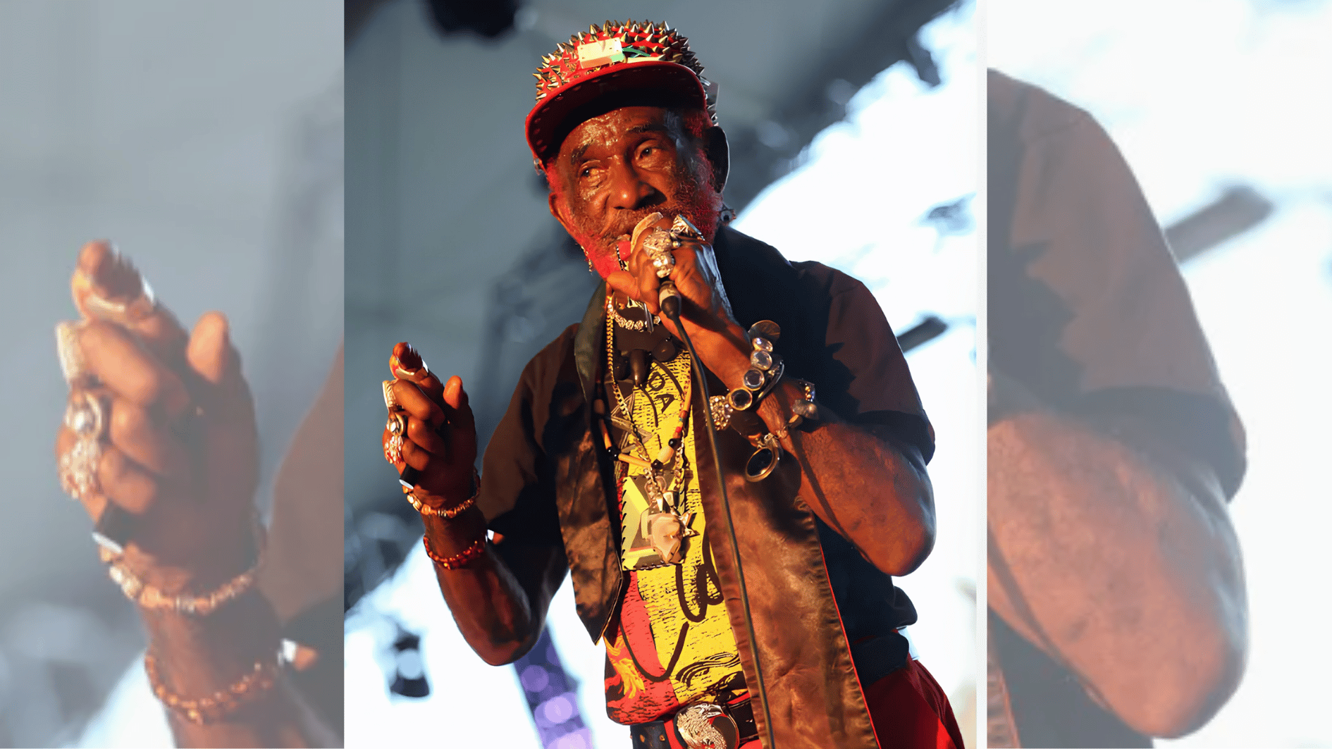 older performer in red pants and spiked cap singing into microphone under stage lights.
