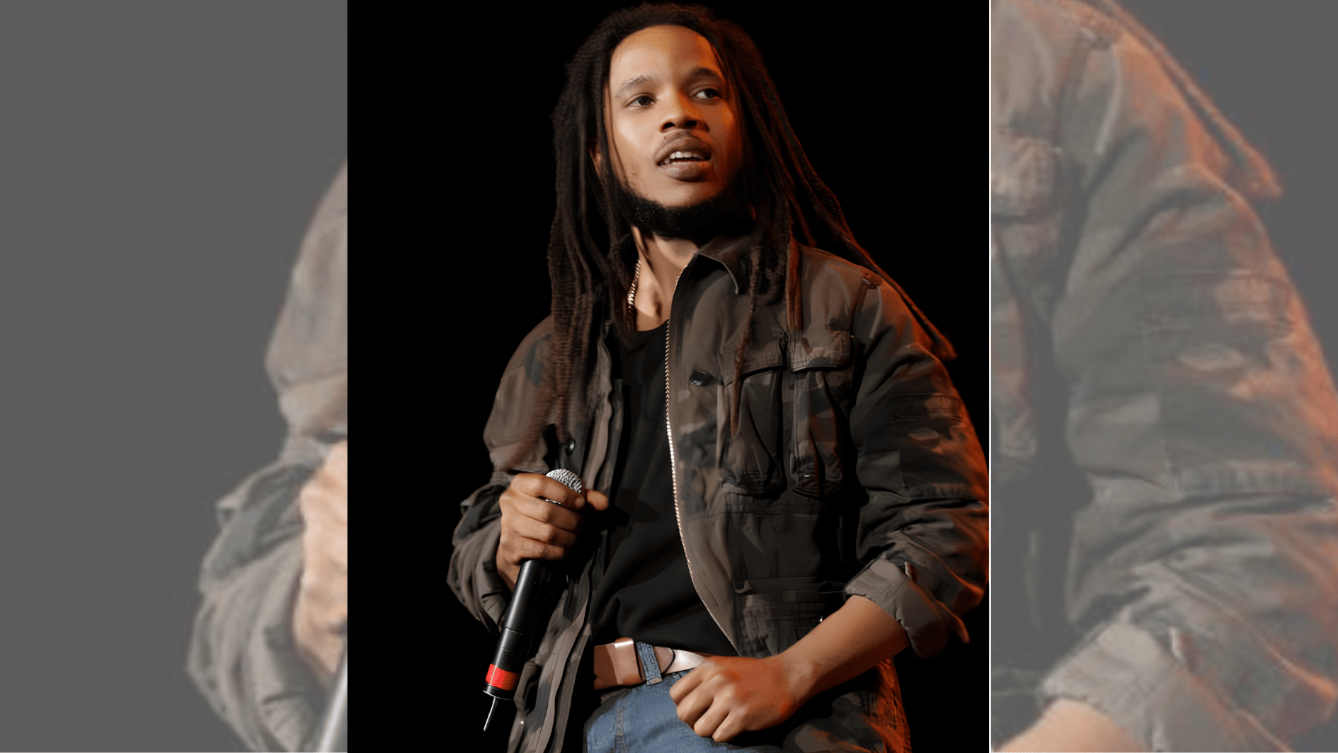 performer with dreadlocks holding microphone while wearing dark jacket onstage.