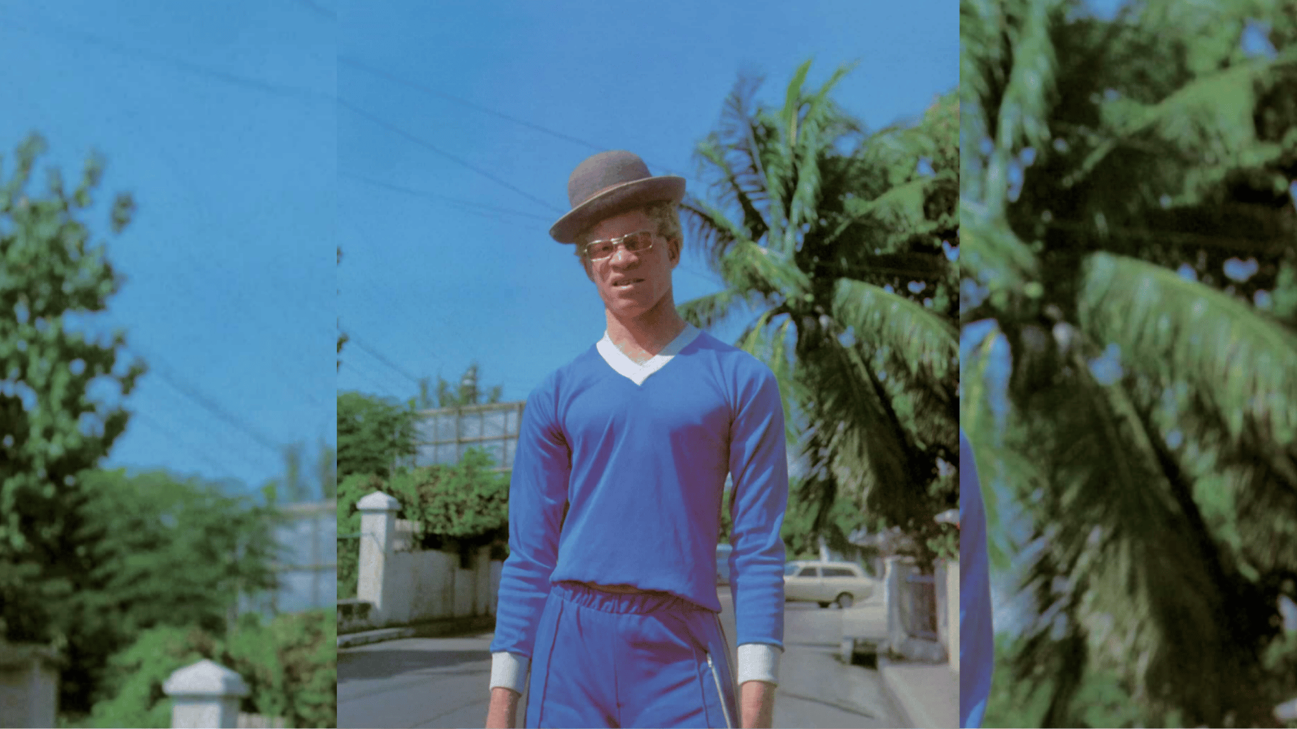 person in blue tracksuit and bowler hat standing on quiet street lined with palm trees.
