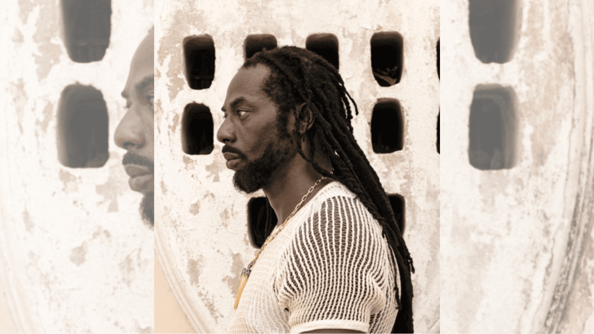 side profile of man with long dreadlocks in a mesh shirt beside a patterned stone wall.